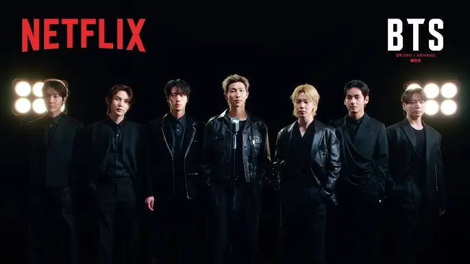 BTS 멤버들이 블랙 수트를 입고 넷플릭스 로고 아래 어두운 배경에서 조명 아래 카리스마 있게 서 있는 모습, 글로벌 K팝 그룹 분위기 강조