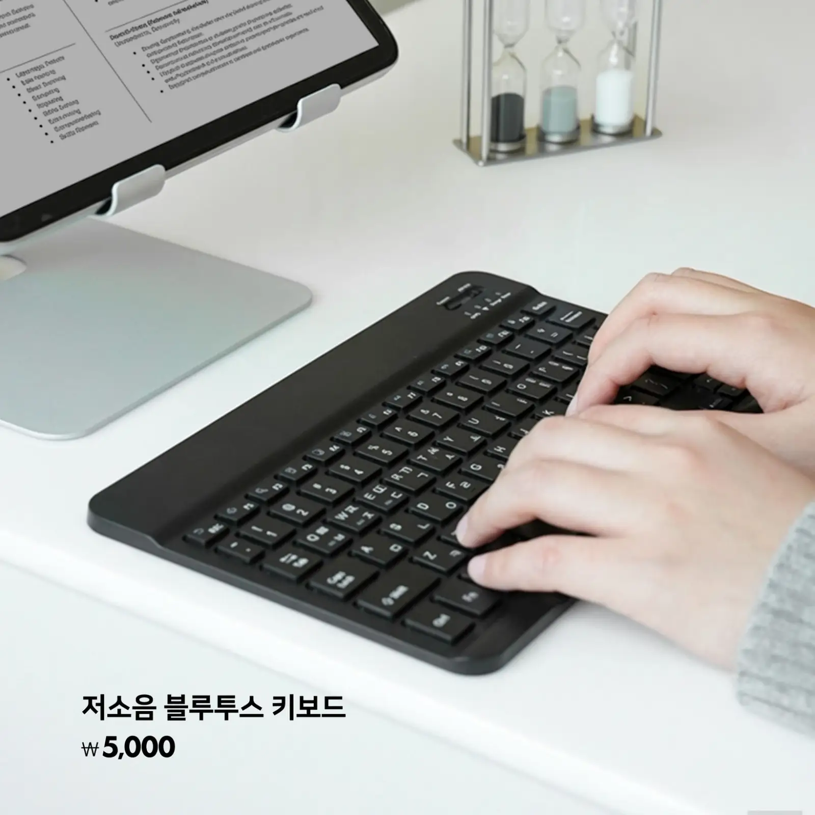 저소음 블루투스 키보드로 깔끔한 책상 위에서 타이핑하는 손, 모던한 사무실 분위기와 컴퓨터, 모래시계 소품이 보임