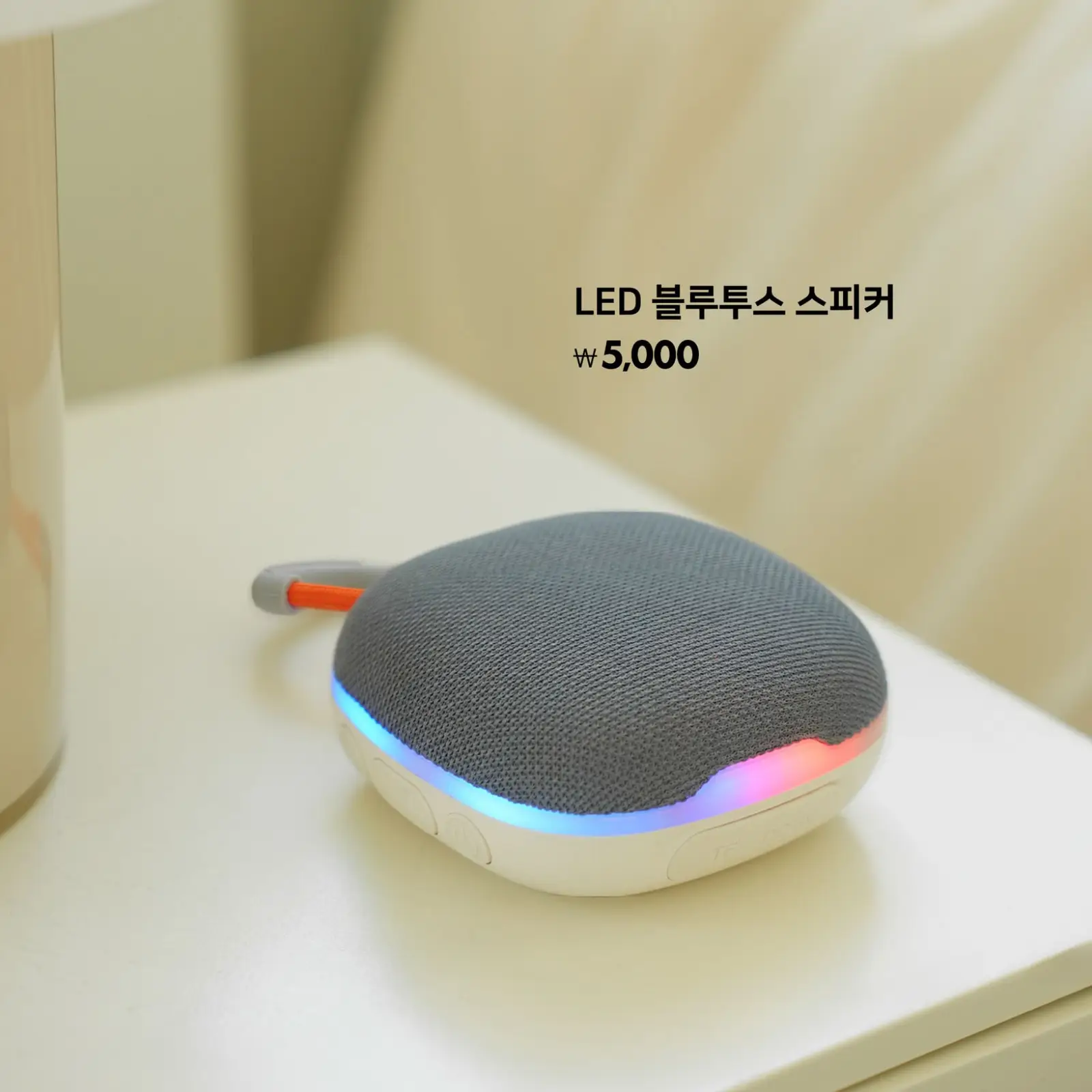 키워드: LED, 블루투스 스피커, 무선, 인테리어, 저가 세련된 그레이 컬러 LED 무드등이 빛나는 무선 블루투스 스피커가 심플한 인테리어 공간 위에 5,000원 가격으로 진열되어 있음.