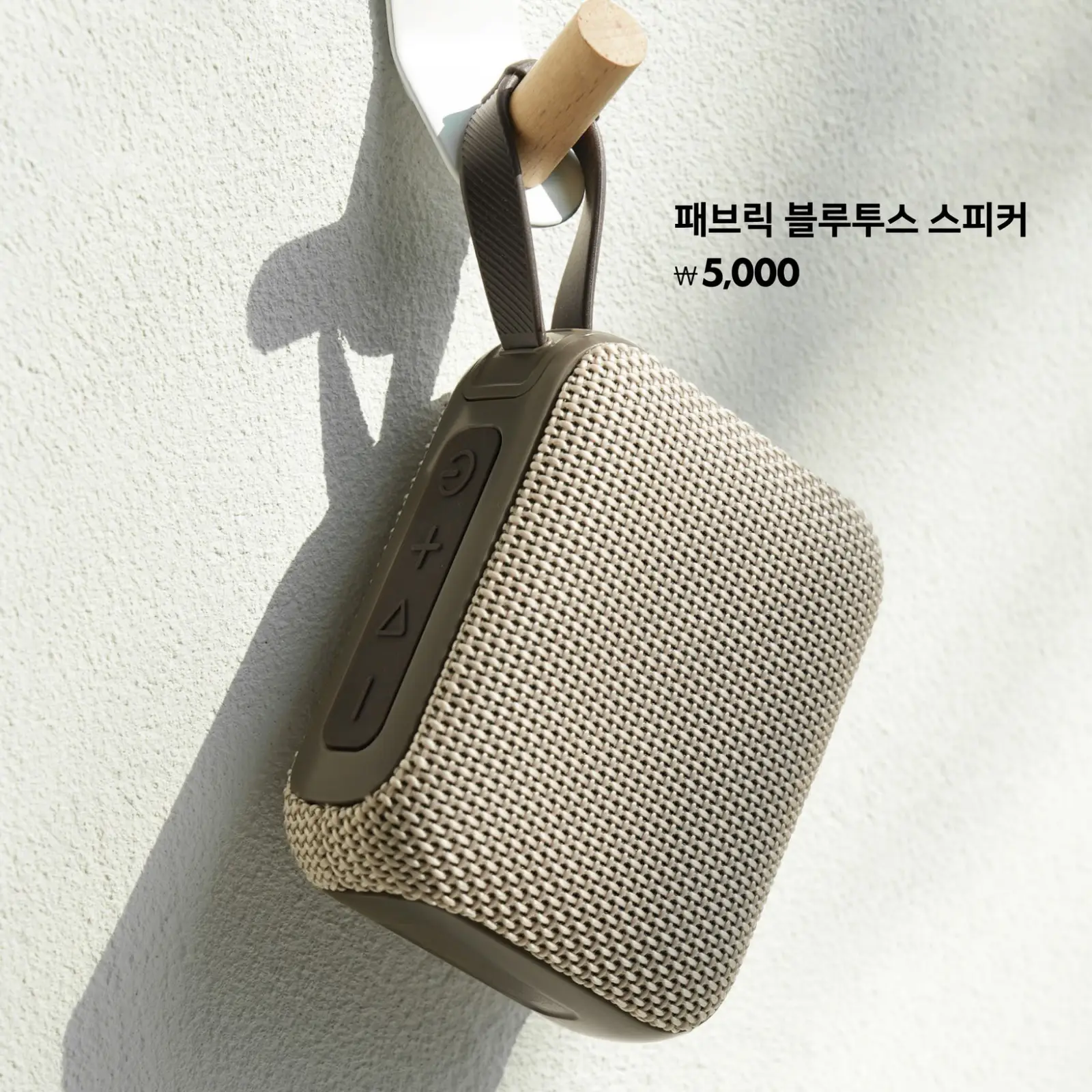 패브릭 블루투스 스피커가 밝은 자연광 아래 벽에 걸려 있으며, 심플한 디자인과 휴대성을 강조한 모습.