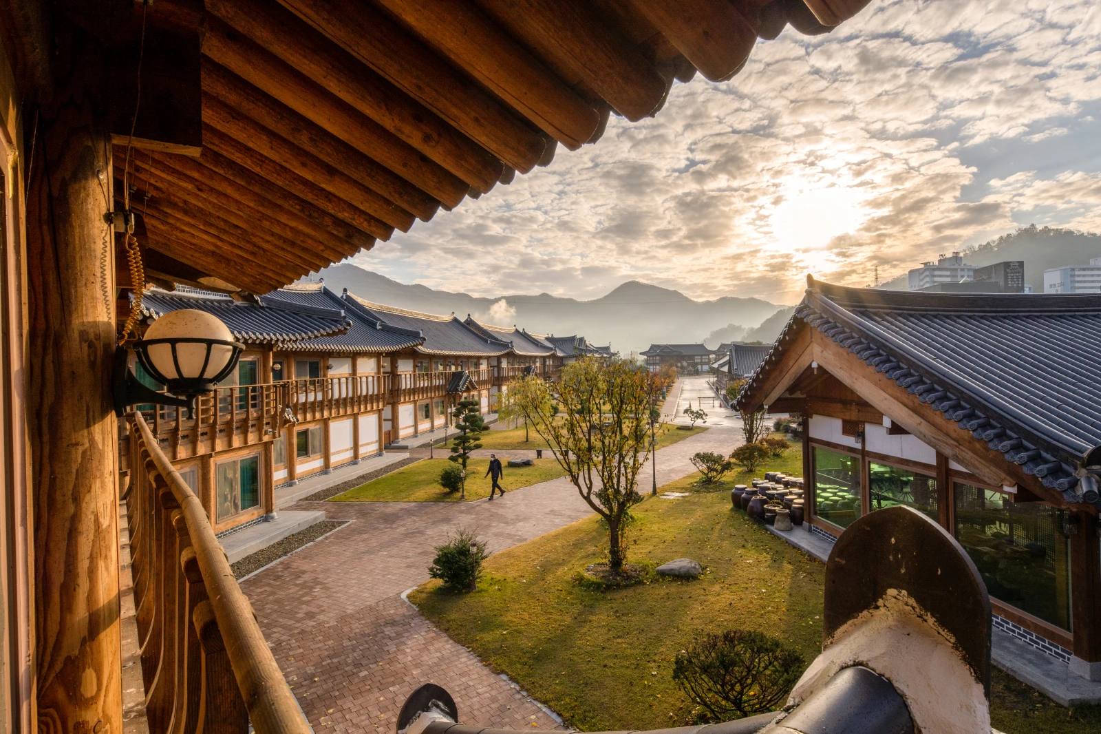 전통 한옥 건물들이 있는 마을 풍경과 뜨는 해, 산과 구름이 배경에 보인다.