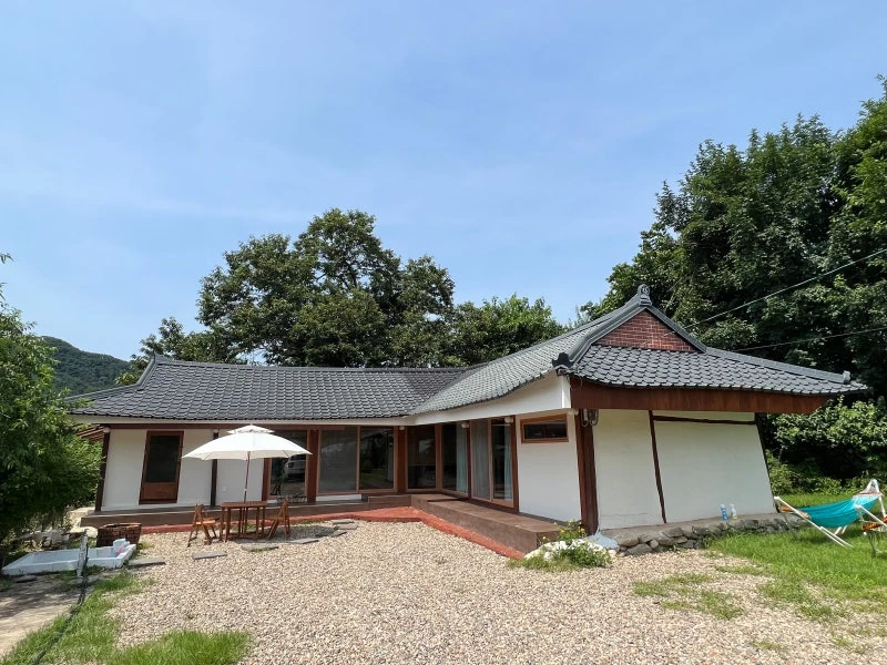 전통 한옥 스타일의 집이 푸른 나무와 하늘 아래에 위치해 있으며 마당에 테이블과 파라솔이 놓여 있다.