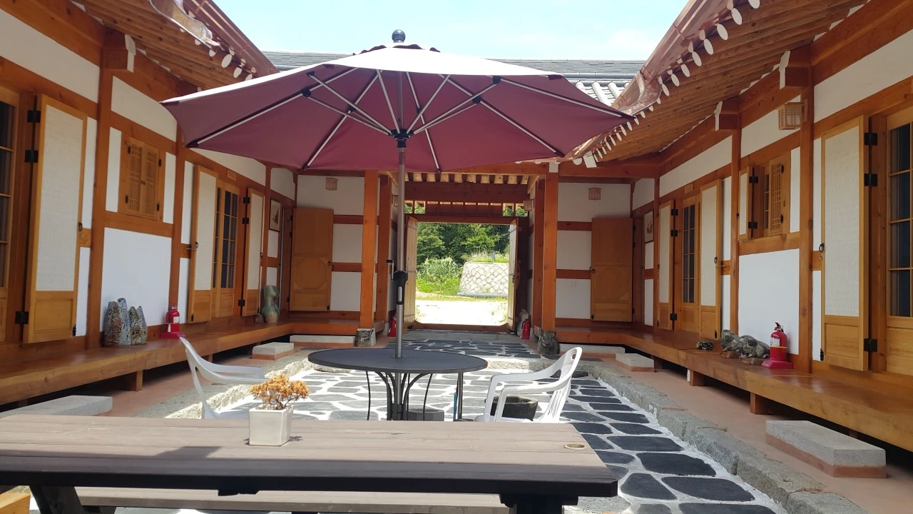 전통 한옥 마당에 큰 파라솔과 테이블, 하얀 플라스틱 의자가 놓여 있고, 나무로 된 건물이 둘러싸고 있다.