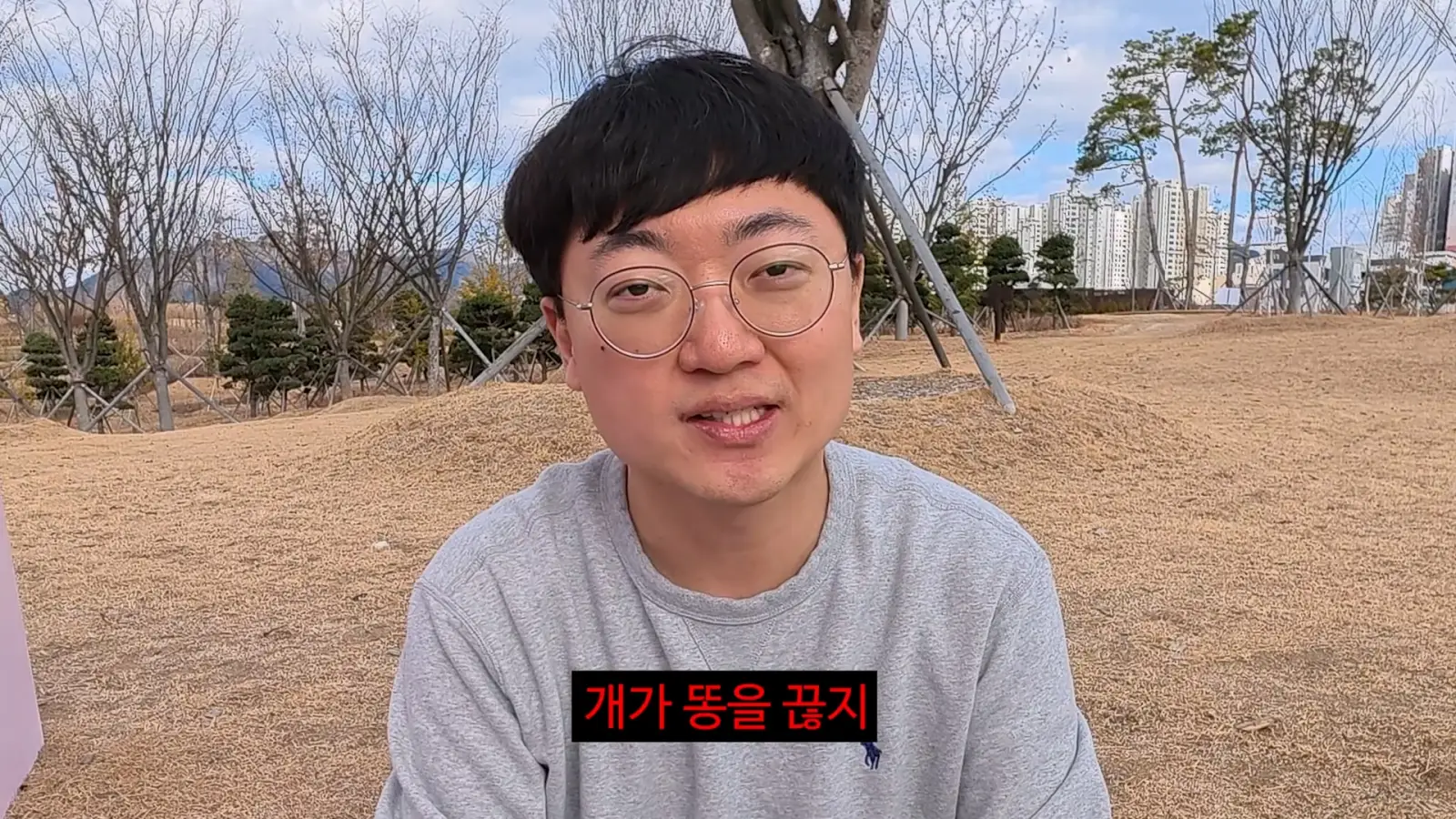안경을 쓴 남성이 공원에서 카메라를 바라보고 있으며, 화면에 "개가 똥을 끊지"라는 자막이 나와 있다.