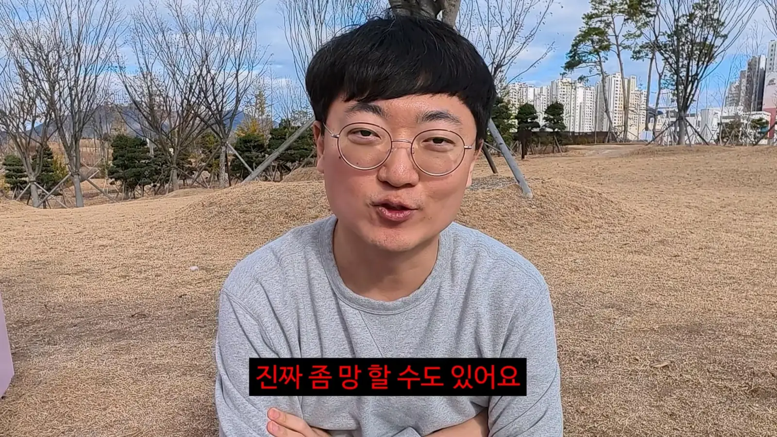안경을 쓴 남성이 회색 티셔츠를 입고 공원에서 팔짱을 끼고 앉아 있다. 화면 아래 자막이 있다.