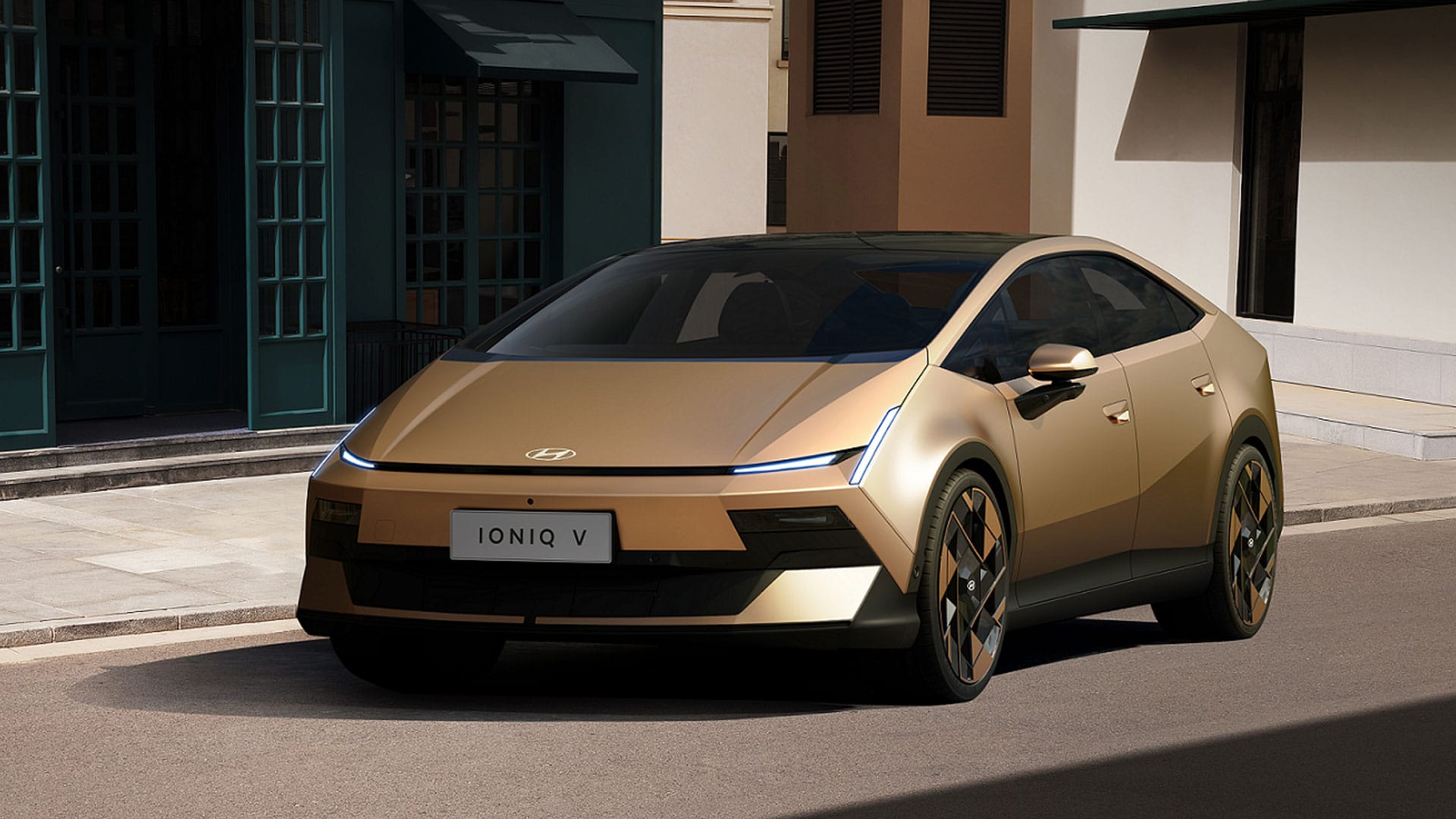 현대 아이오닉 V 전기차의 외관을 보여주는 이미지로, 세련된 디자인과 금색 외장, 독특한 휠이 특징이다. 도시 배경에서 주차된 모습으로, 미래 지향적인 스타일을 강조한다.