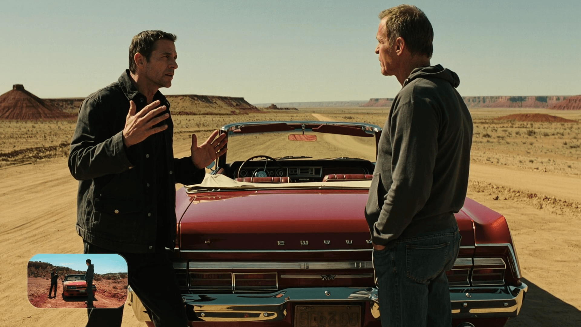 두 남성이 드넓은 사막 배경에서 빨간 클래식 자동차 앞에서 대화하고 있는 장면. 한 남성이 손짓하며 이야기를 하고 있고, 다른 남성은 주의 깊게 듣고 있는 모습.