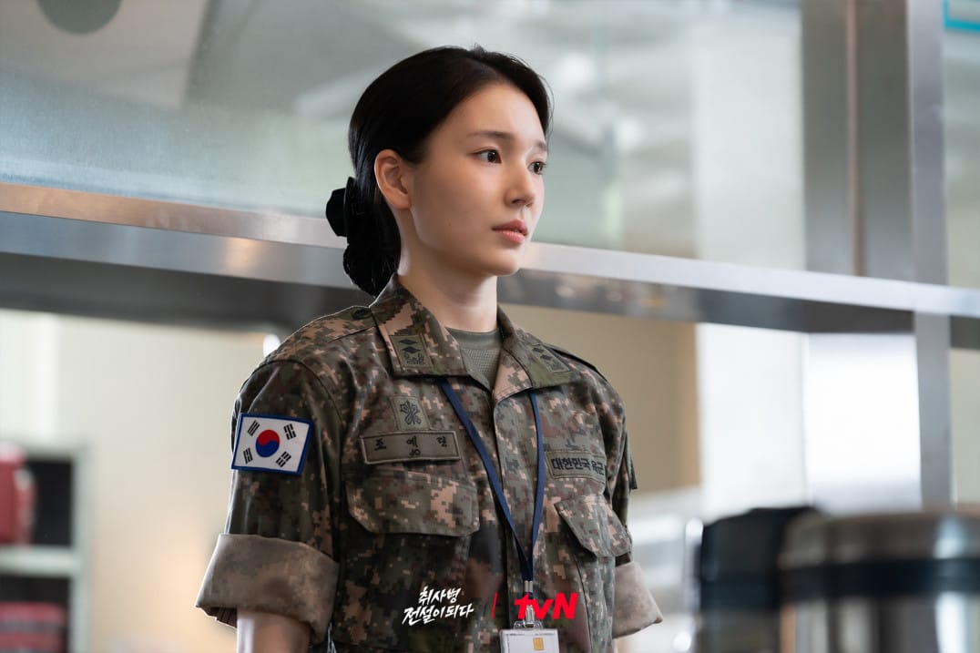 한국 군복을 입고 있는 여성 군인의 모습, 진지한 표정으로 서 있는 장면. 배경에는 군 관련 시설이 보입니다.