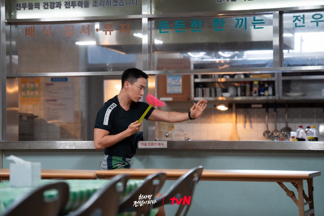주방에서 요리를 준비하는 남성이 분주하게 움직이고 있는 모습, 손에 식기 세척 도구를 들고 있으며, 배경에는 식당 내부가 보인다.