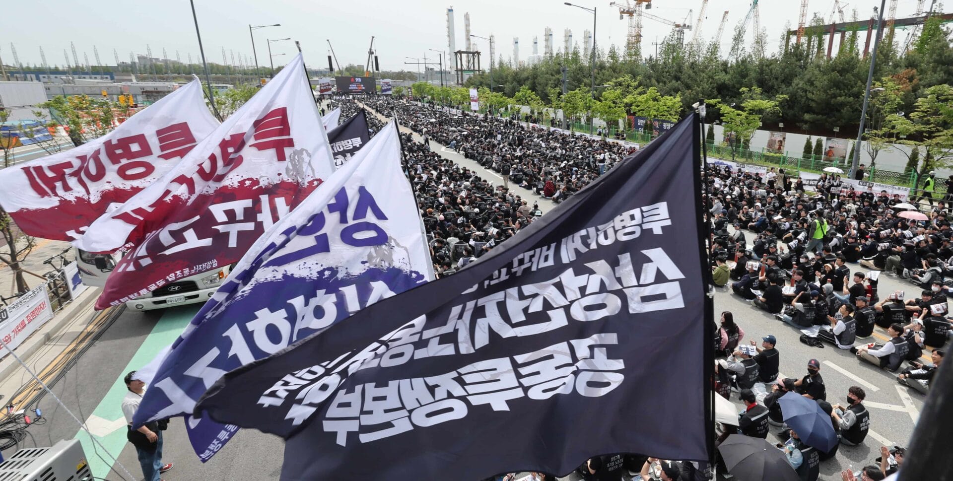 대규모 시위에서 다양한 깃발이 휘날리며 많은 사람들이 도로에 앉아 있는 모습. 시위 참가자들은 검은색 의상을 입고 있으며, 주변에는 건설 중인 구조물과 나무가 보인다.