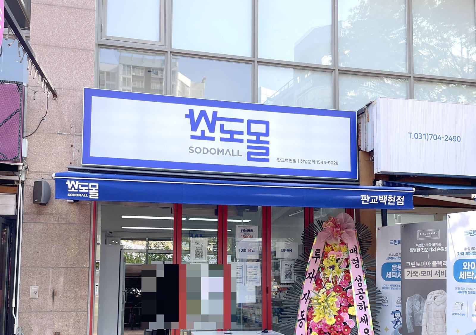 상점 외관 사진으로, '쏘도몰'이라는 간판이 보이며, 주변에 꽃다발과 다양한 광고 배너가 위치해 있습니다. 상점은 현대적인 디자인으로 창문이 크고 밝은 분위기를 자아내고 있습니다.