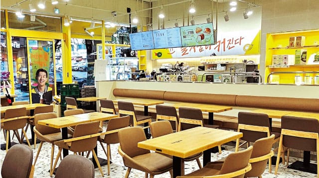 카페 내부 모습, 현대적인 디자인의 테이블과 의자가 배치되어 있으며, 밝은 노란색 벽면과 메뉴판이 눈에 띄는 공간.