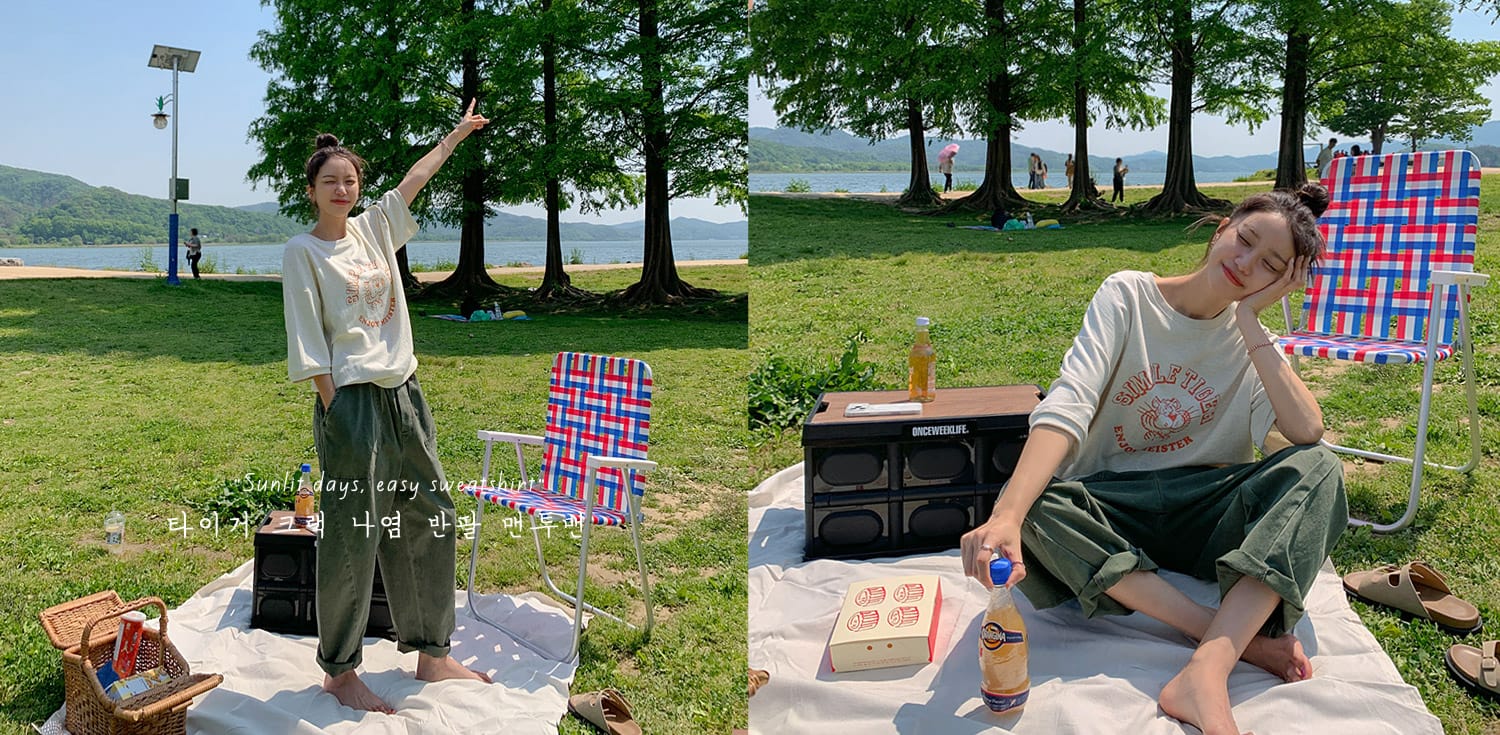 햇살 가득한 날, 수변에서 편안한 옷을 입고 앉아 있는 여성. 푸른 잔디와 나무 아래에서 음료수와 간식을 즐기며 여유로운 시간을 보내는 모습. 배경에는 호수와 산이 보임.