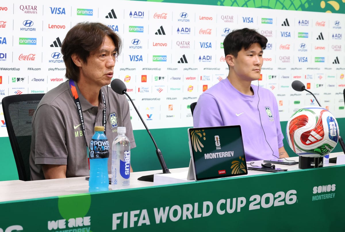 2026 FIFA 월드컵을 앞두고 기자회견에 참석한 한국 축구 감독과 선수의 모습. 감독은 마이크 앞에서 발언 중이며, 선수는 옆에서 경청하고 있다. 테이블 위에는 축구공과 전자기기가 놓여 있다. 배경에는 다양한 스폰서 로고가 보인다.