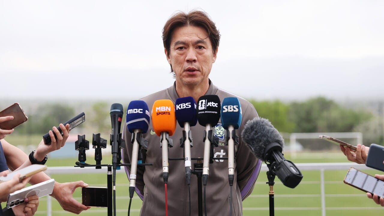 축구 감독이 기자회견 중 마이크 앞에 서 있는 모습, 다양한 미디어 장비와 기자들이 질문을 준비하고 있는 장면.