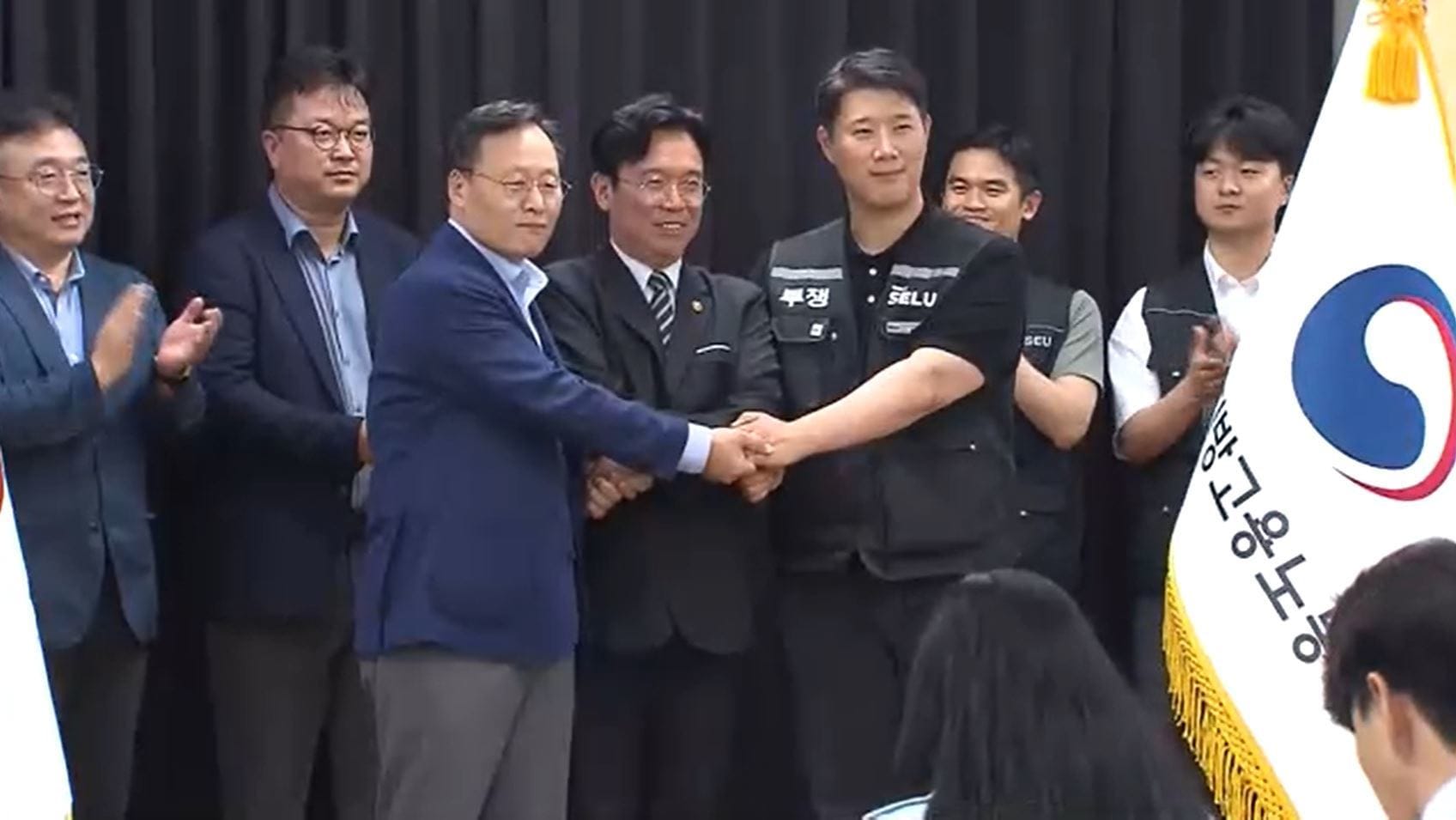 노동 관련 행사에서 여러 인사들이 손을 맞잡고 있는 모습. 왼쪽에는 박수를 치고 있는 참석자들이 있으며, 배경에는 한국의 국기가 보인다.