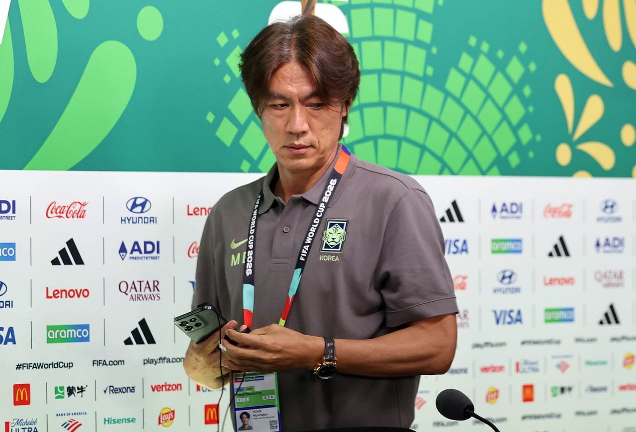 한국 축구 국가대표팀 감독이 FIFA 월드컵 2026 관련 기자회견 준비 중인 모습, 스마트폰을 들고 있으며, 팀 유니폼을 착용하고 있다. 배경에는 여러 스폰서 로고가 보인다.
