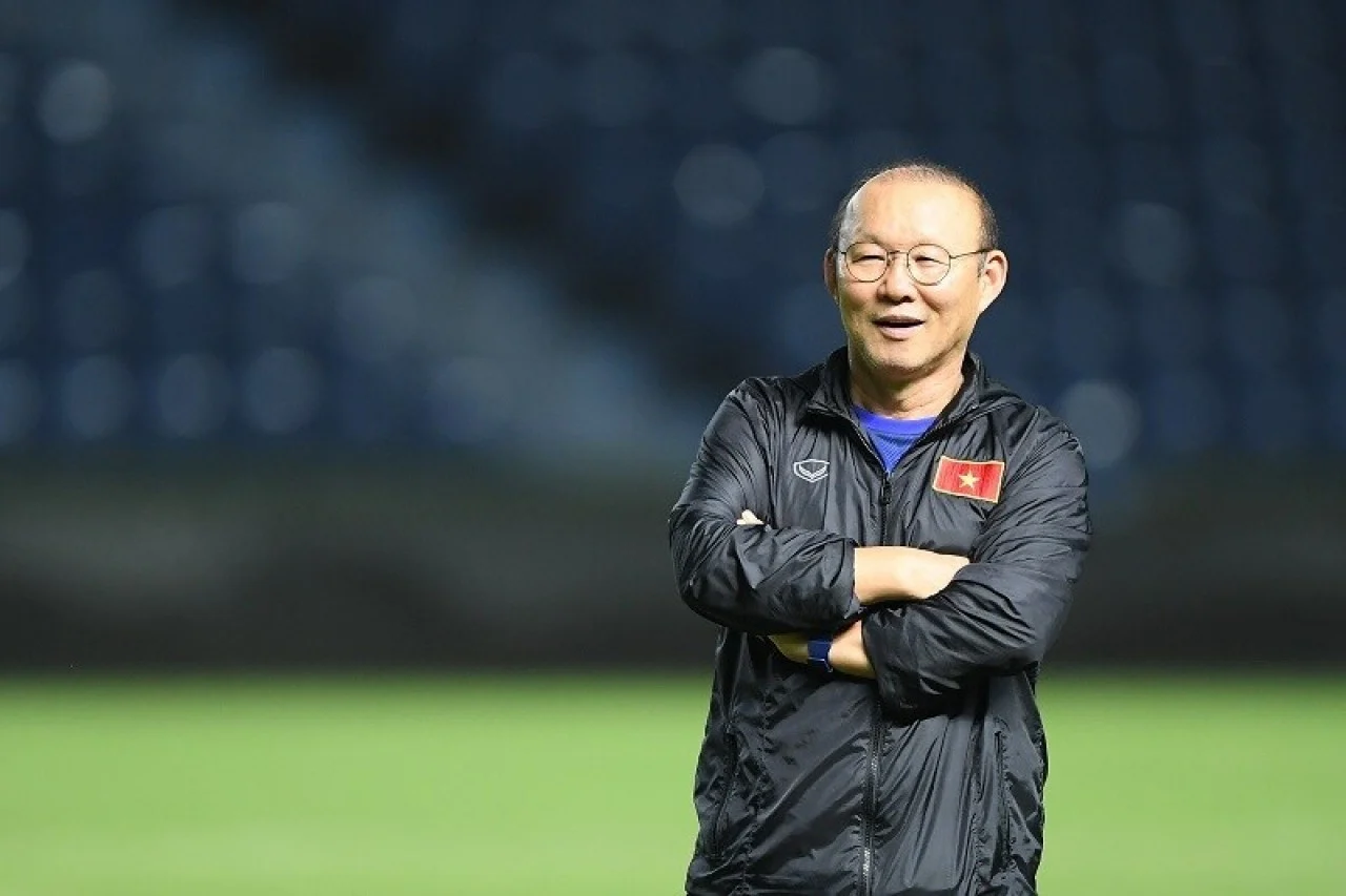 축구 훈련장에서 미소 짓고 있는 베트남 축구 감독, 검은색 재킷을 입고 있으며, 베트남 국기가 부착되어 있는 모습.
