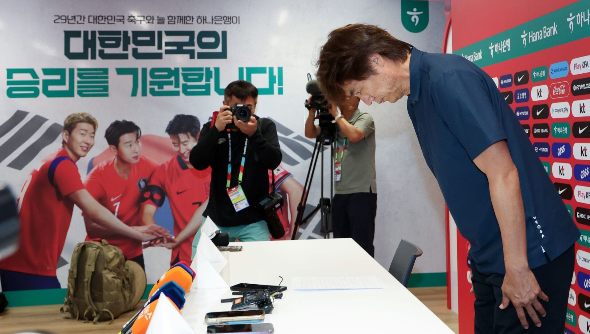 축구 기자회견에서 고개를 숙이고 있는 감독과 카메라를 향해 촬영하는 기자들, 배경에는 한국 축구 대표팀의 응원 메시지가 담긴 포스터가 보입니다.