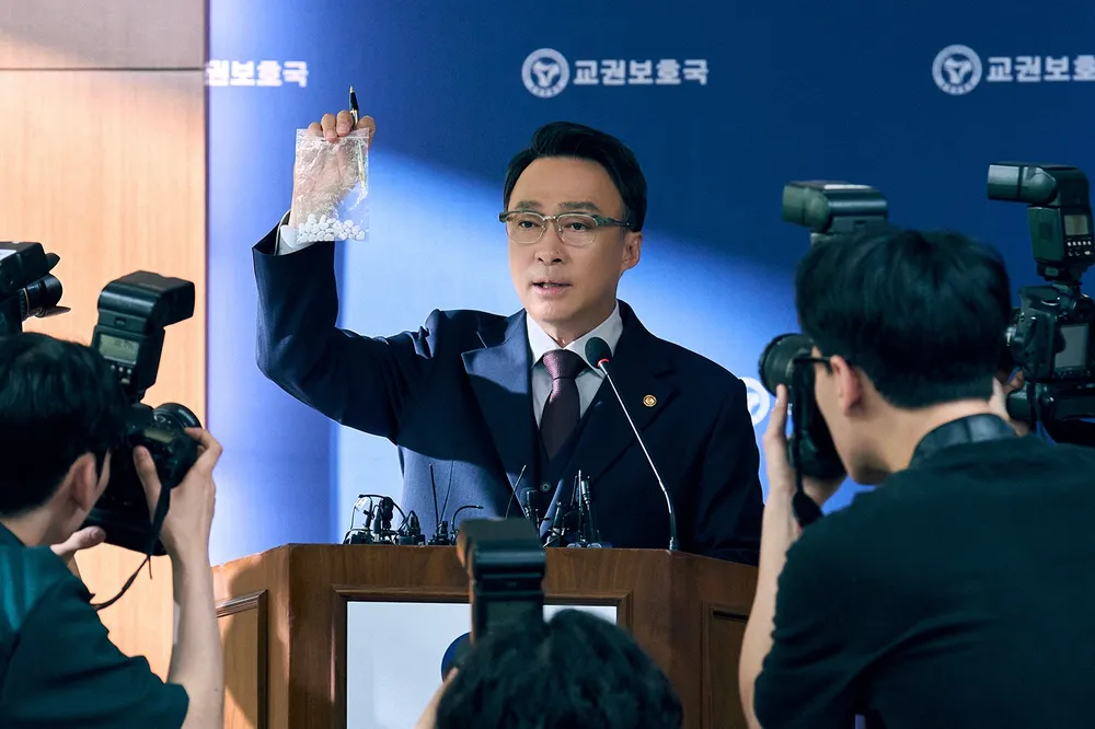 교권 보호국의 공식 기자회견에서 한 남성이 투명한 봉투에 담긴 알약을 들어 보이며 발언하고 있는 모습, 주변에는 여러 사진 기자들이 집중하고 있음.