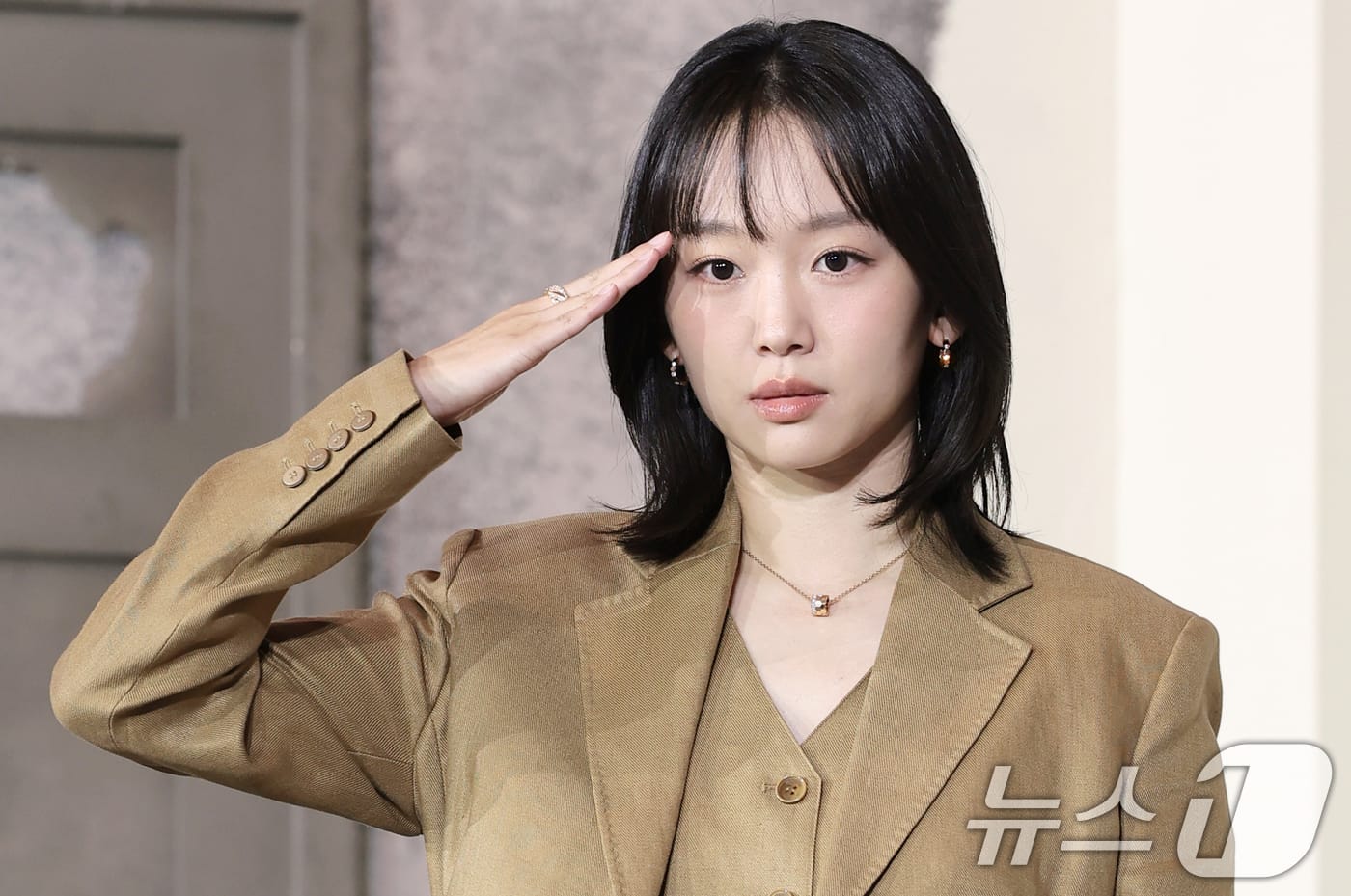 여성 모델이 갈색 정장을 입고 경례하는 모습. 그녀는 짧은 머리 스타일과 심플한 액세서리를 착용하고 있으며, 배경은 흐릿하게 처리되어 있다.