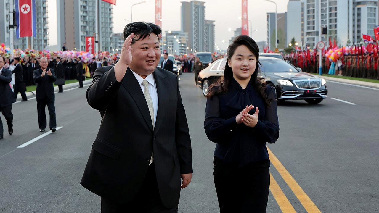 김정은과 그의 딸이 군중 앞에서 손을 흔들며 걷고 있는 모습, 뒤에는 차량과 북조선 국기가 보임.