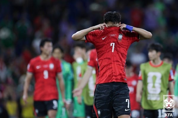 손흥민이 경기 후 실망한 모습으로 셔츠를 들어올리고 있는 장면, 배경에는 한국 축구팀 선수들이 보인다. 이 이미지는 손흥민의 선발 제외 이유와 관련된 논란을 다루고 있는 기사에 사용됨.