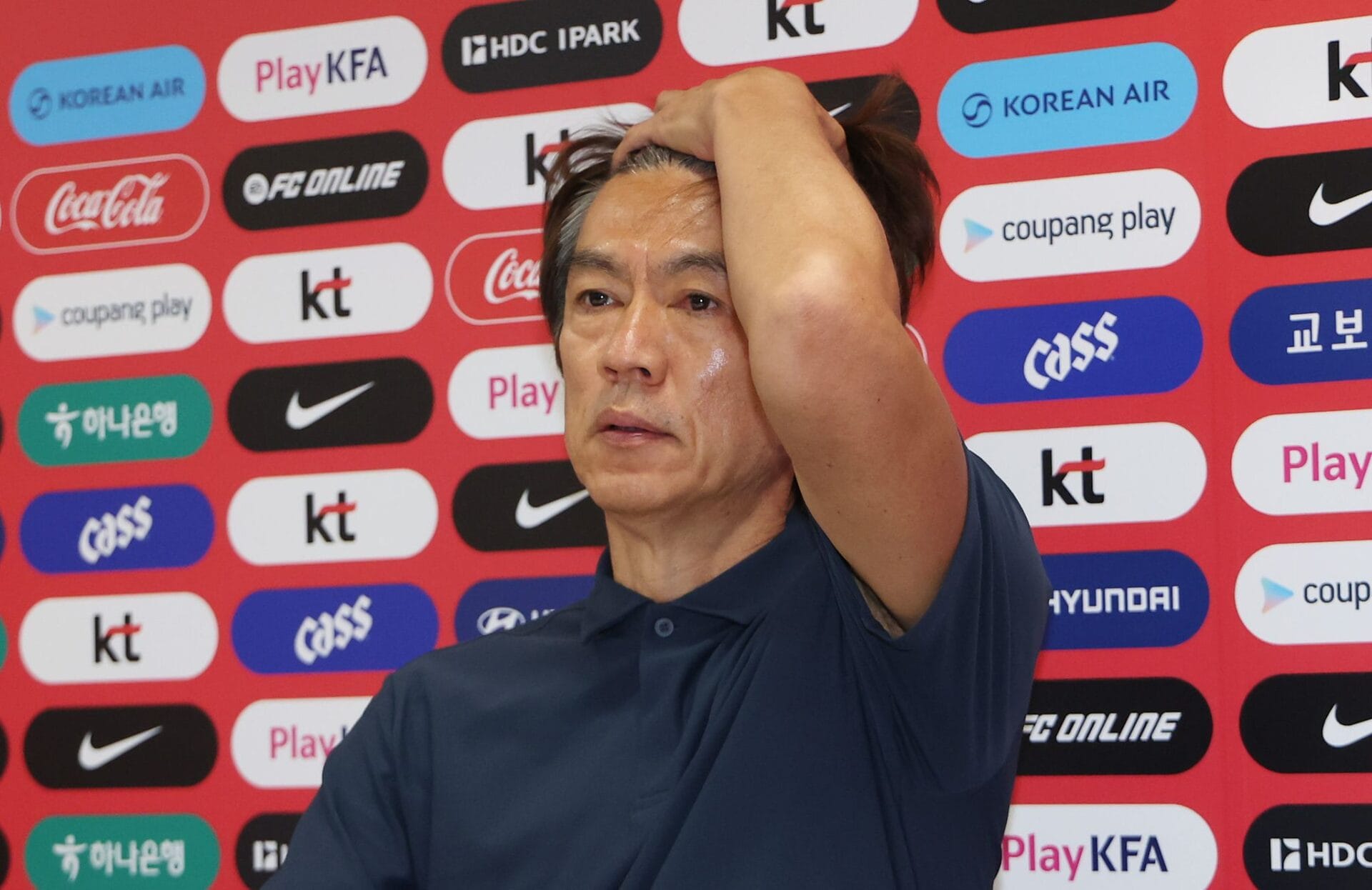 손흥민 선발 제외에 대한 인터뷰를 거부한 감독의 모습, 긴장한 표정과 함께 머리를 감싸고 있는 모습이 포착됨. 배경에는 다양한 스폰서 로고가 있는 기자회견장.