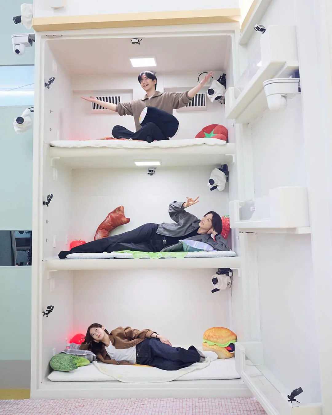 대환장 기안장 시즌2의 출연자들이 독특한 침대 공간에서 다양한 포즈를 취하고 있는 모습, 각 침대에는 음식 모양의 인형들이 배치되어 있으며, 카메라와 조명이 설치된 현대적인 환경을 보여줌.