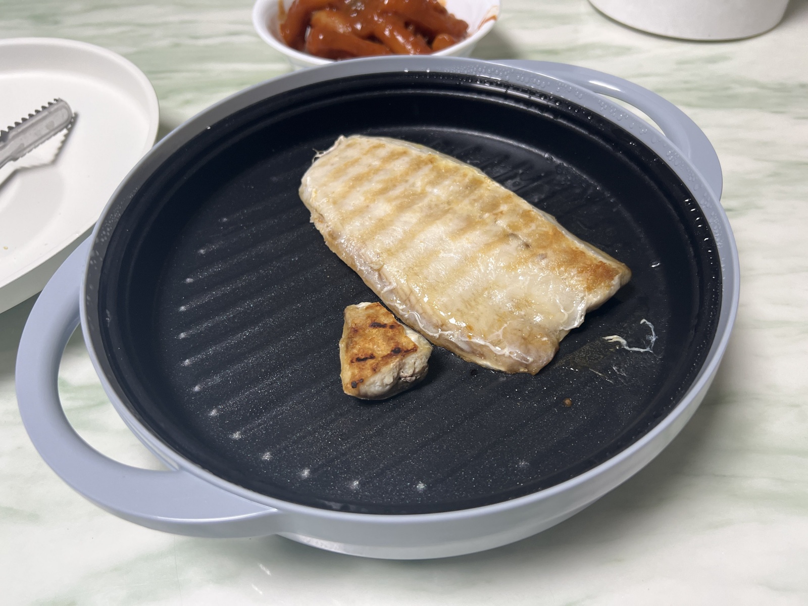전자레인지에서 조리된 고등어구이가 담긴 그릴 팬, 부드러운 식감과 맛있는 갈색 크러스트가 특징이며, 옆에는 작은 고기 조각이 함께 놓여 있음.