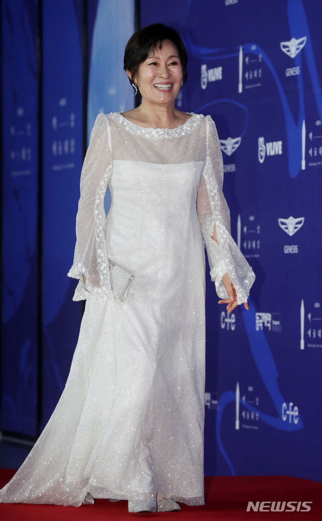 부산국제영화제에서 화려한 드레스를 입고 미소 짓고 있는 김혜자, 길게 늘어진 소매와 반짝이는 소재의 드레스가 돋보임.